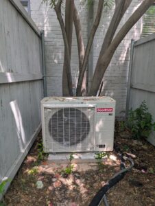 Here are three options for describing this image: Alt Text: "Goodman air conditioning unit sitting on concrete pad in backyard corner between wooden fences,