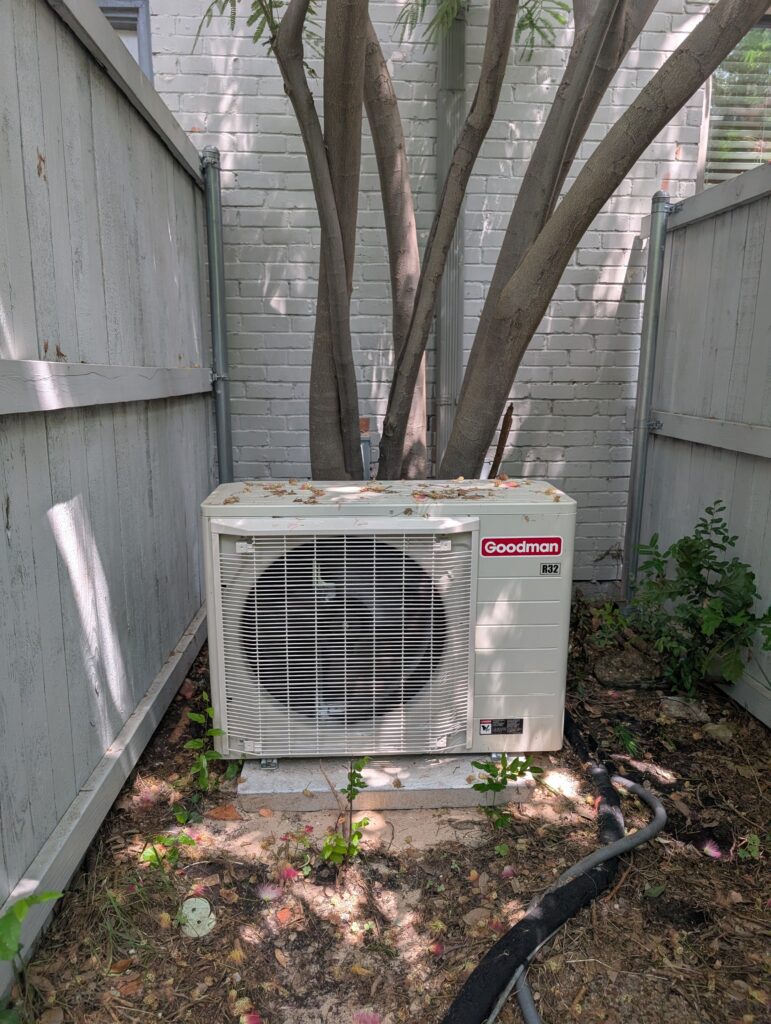 Here are three options for describing this image:
Alt Text:
"Goodman air conditioning unit sitting on concrete pad in backyard corner between wooden fences,